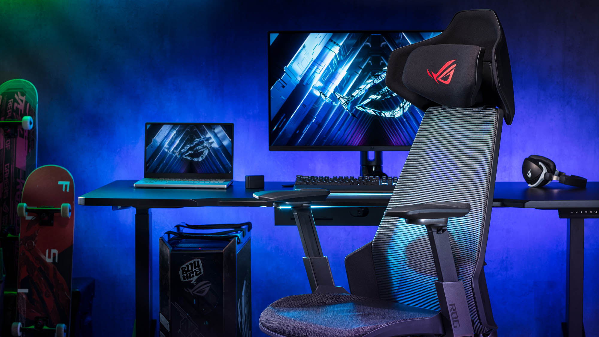 An ROG laptop and desktop setup alongside a pair of ROG headphones, and ROG chair, and skateboards against a purple wall.